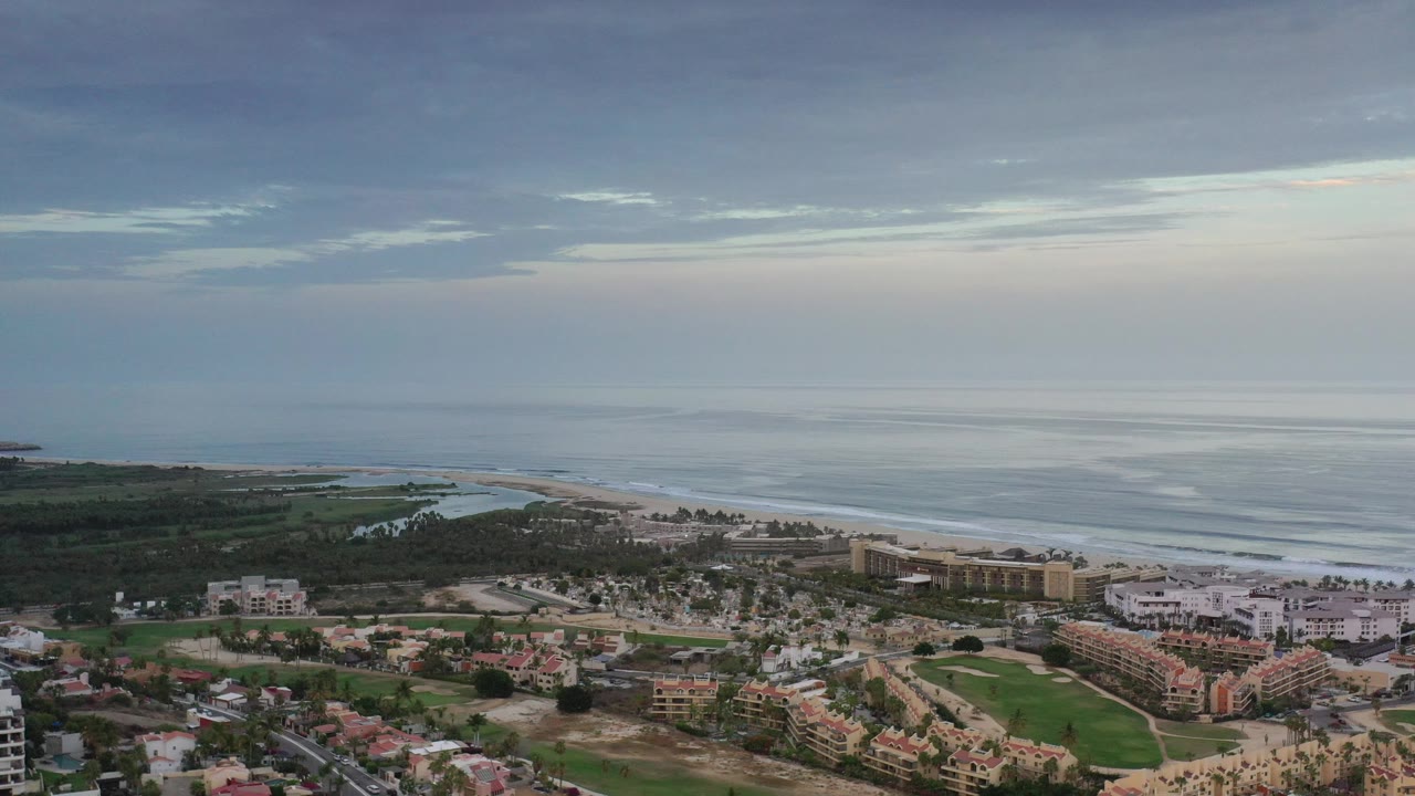 Wide aerial coastline with beachfront buildings, palms, and soft sunset light.