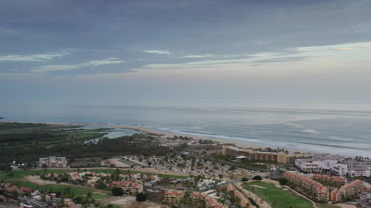 Aerial Los Cabos frame showing the ocean horizon high in the background, pale surf bands along the beach, clustered buildings below, and no visible people.