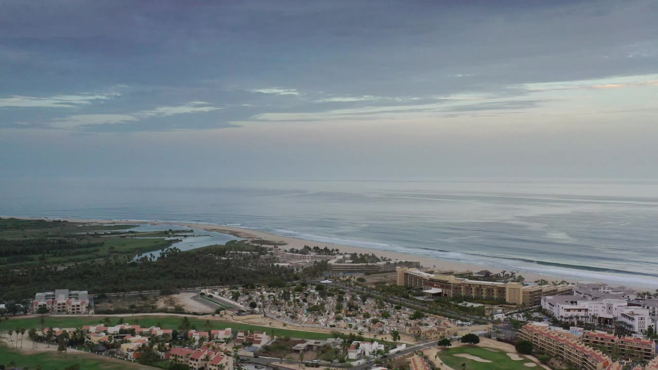 High Los Cabos coastal view with hotel blocks near the sand, neighborhood rooftops below, a curving inlet to the left, and no visible people.