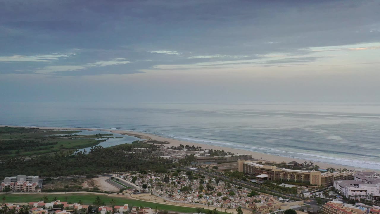 Los Cabos aerial frame showing the ocean stretching across the background, a pale beach curve, green tree cover, and resort-scale buildings near the lower right, with no visible people.
