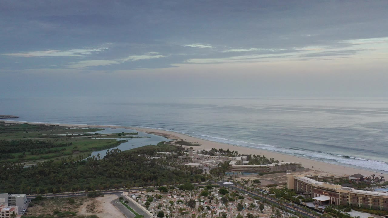 Opening aerial frame over Los Cabos, Mexico with green lagoon water at left, a pale beach and surf line at center, low buildings below, and no visible people.