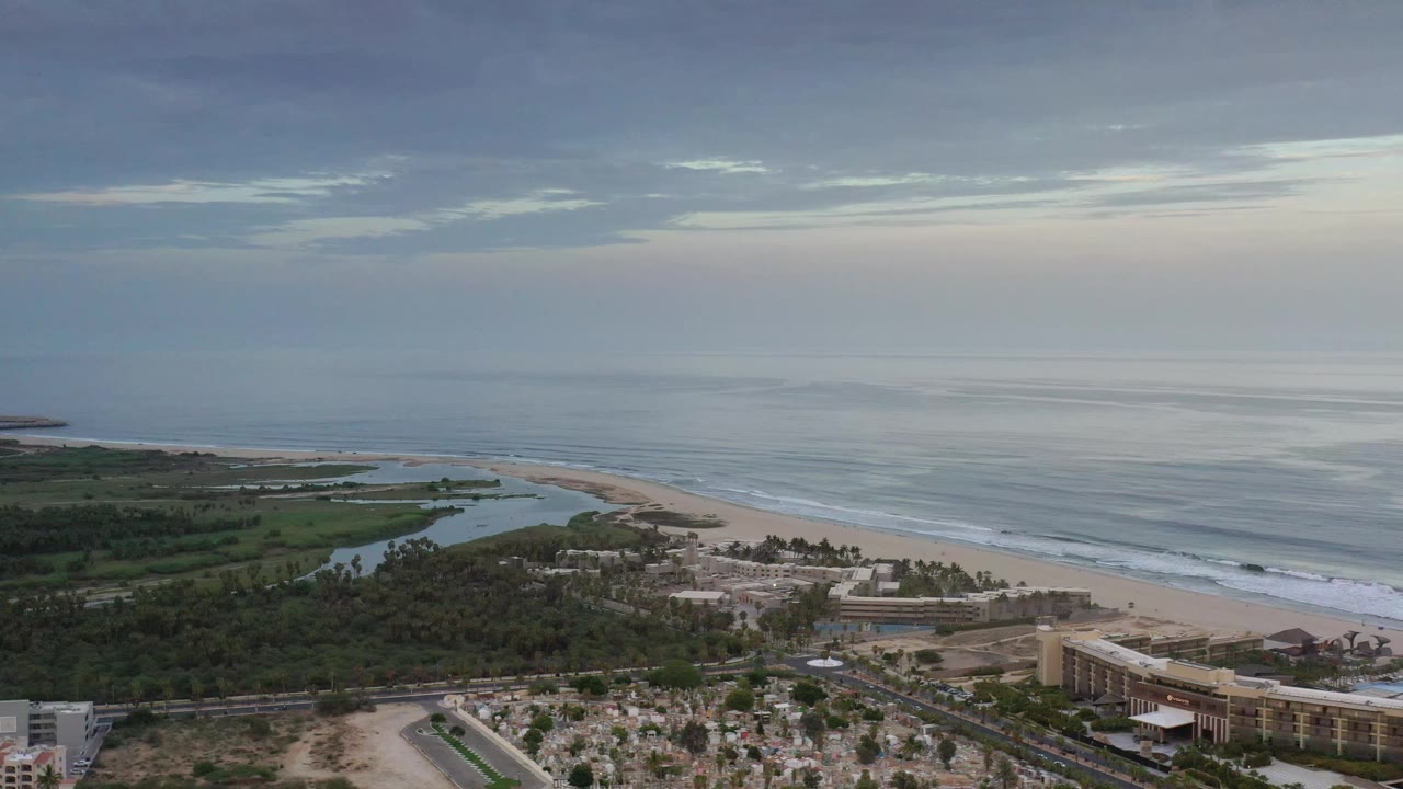 Aerial view in Los Cabos, Mexico with the shoreline running diagonally across the frame, lagoon water behind the beach, buildings along the lower edge, and no visible people.