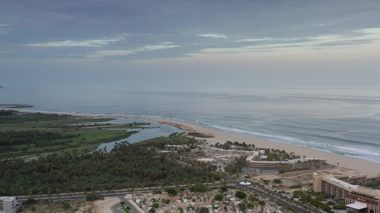 Opening aerial frame over Los Cabos, Mexico with dense beachside trees, a pale sandy shoreline, calm ocean bands, a cloudy blue-gray sky, and no visible people.