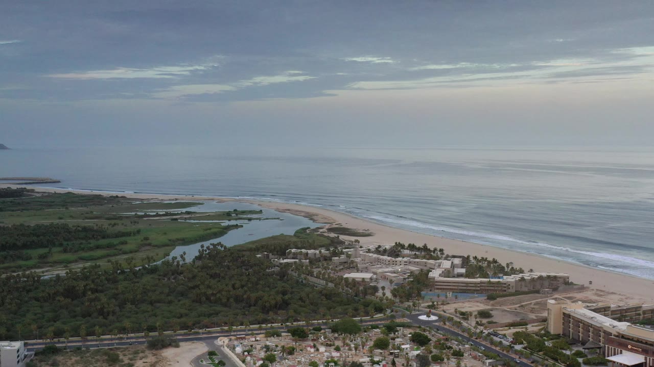 Wide coastal landscape with ocean horizon, sandy beach, trees, and resort-scale buildings.