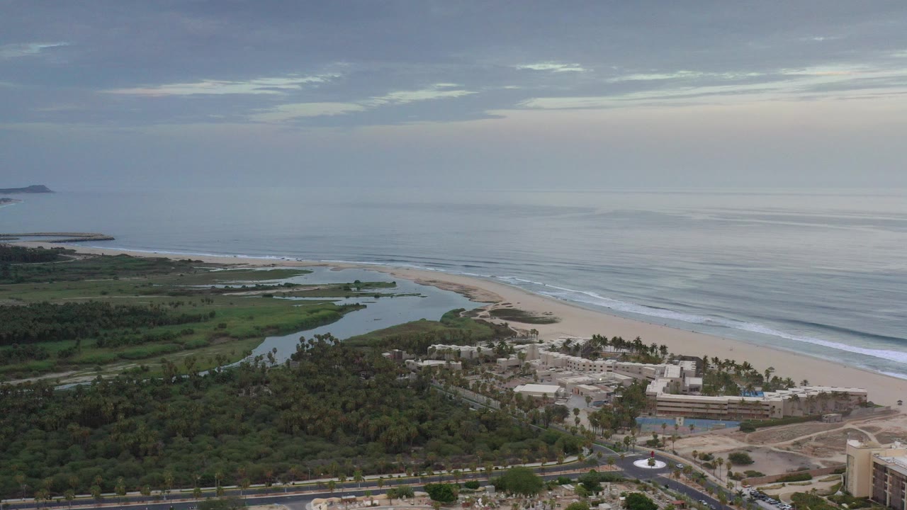 Wide aerial view over Los Cabos, Mexico, showing green trees, a lagoon, sandy beach, ocean surf, and no visible people at the start of the clip.