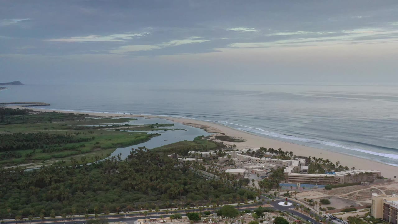 Aerial frame over Los Cabos, Mexico, with the lagoon curving toward the beach, pale surf bands offshore, treetops below, and no visible people.