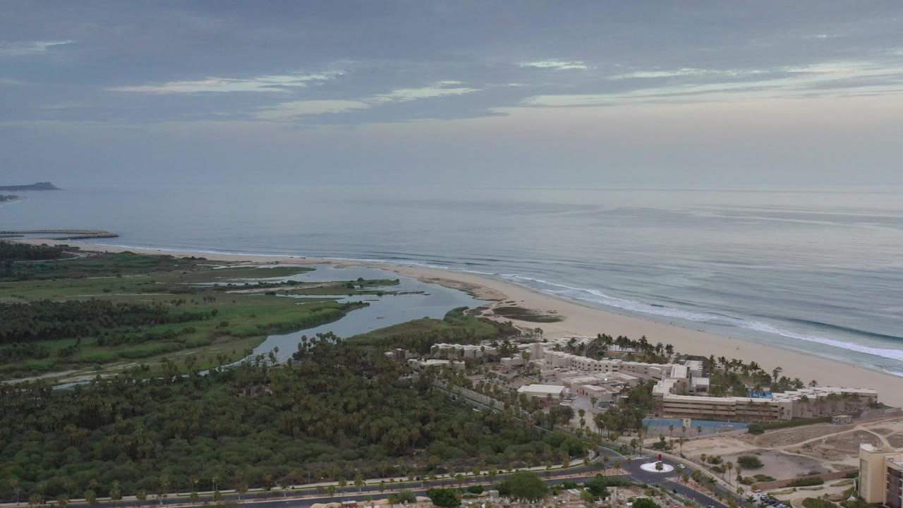 Coastal aerial with ocean, beach, lagoon, trees, and resort-like buildings below.