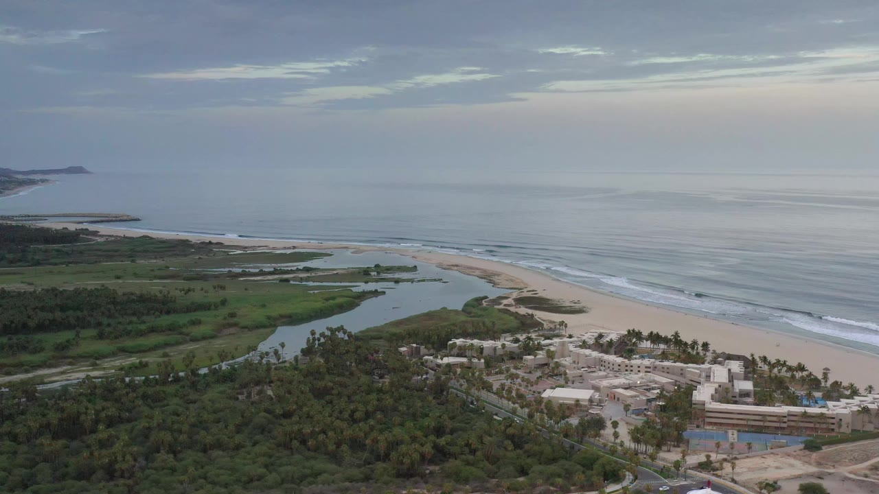 Aerial frame in Los Cabos, Mexico, showing the inlet curving through green land toward a wide sand beach and muted ocean, with low buildings and palms in the foreground and no visible people.