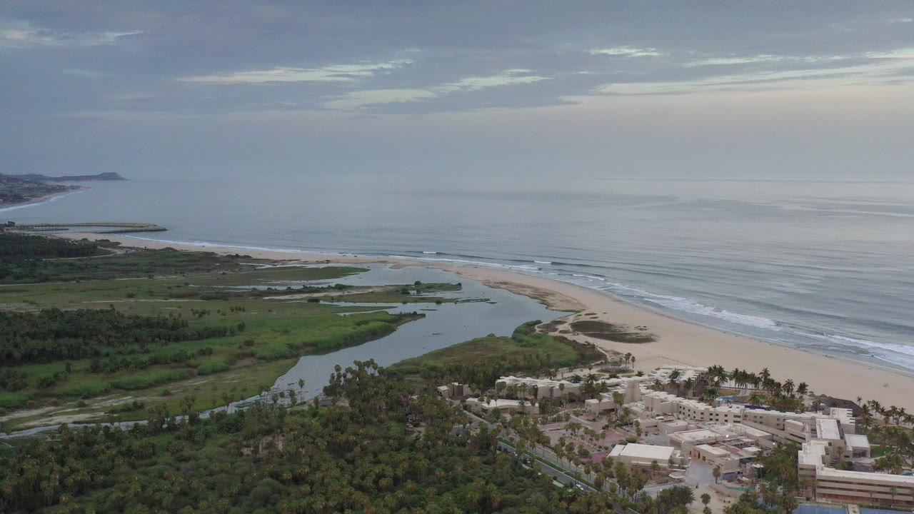 Opening aerial frame over Los Cabos, Mexico, showing low-rise coastal buildings in the foreground, a lagoon behind the beach, calm ocean beyond, and no visible people.