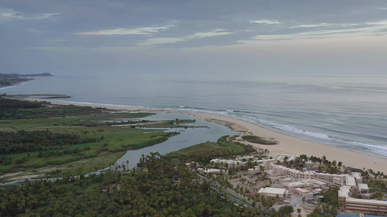 Los Cabos aerial view with the lagoon and greenery more centered, surf lines along the sand, distant coastline on the left, and no visible people.