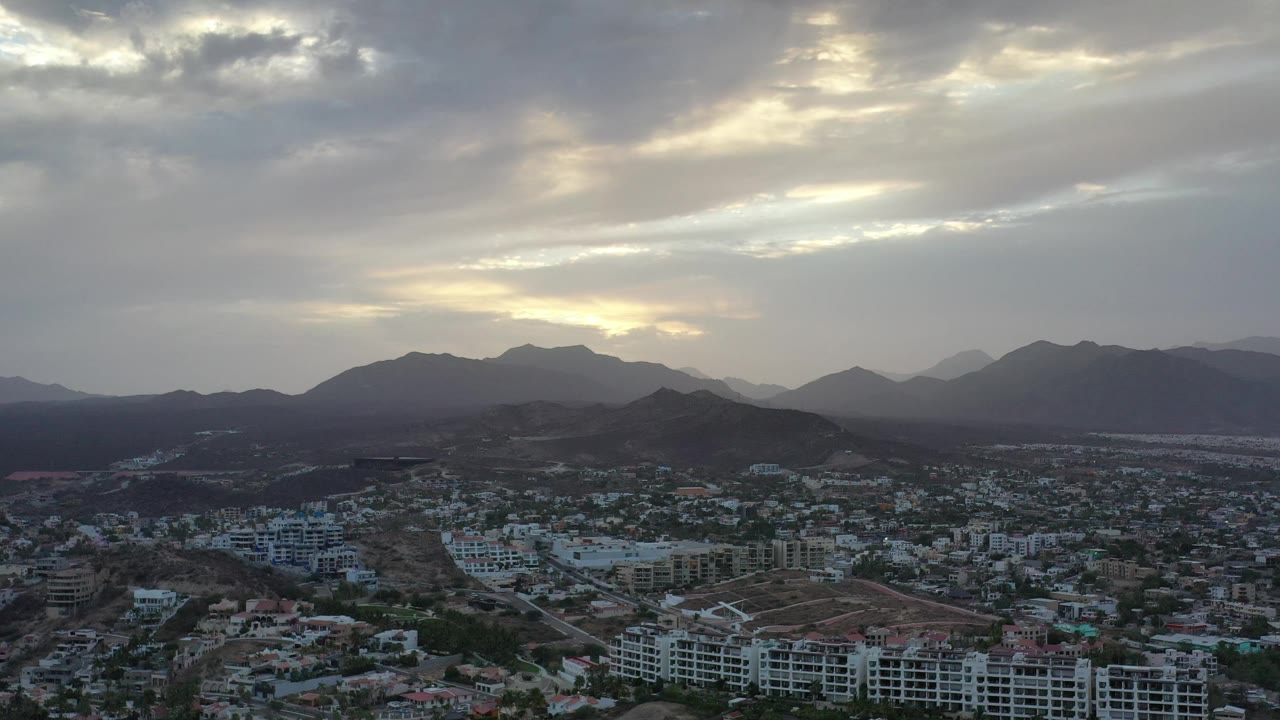 Los Cabos, Mexico aerial view with dense low-rise development in the foreground, darker hills across the middle distance, layered mountains beyond, cloud breaks overhead, and no visible people.