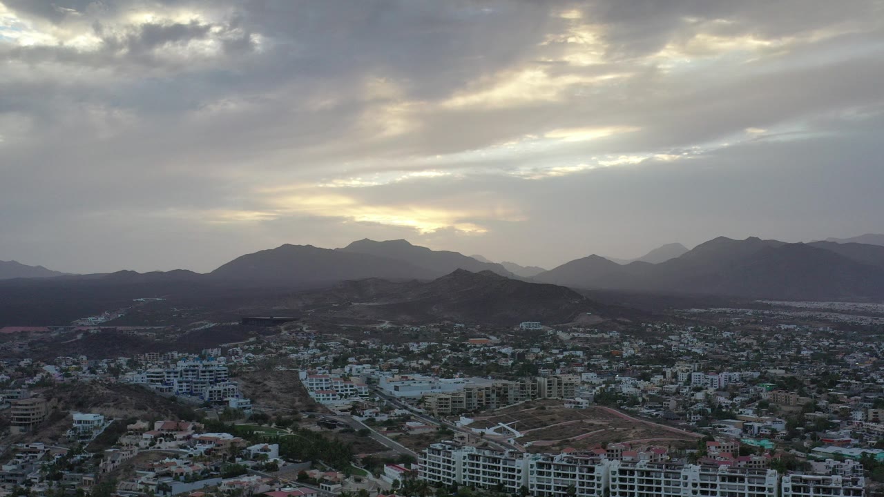 Wide aerial view of a city below mountains and a cloudy sky.