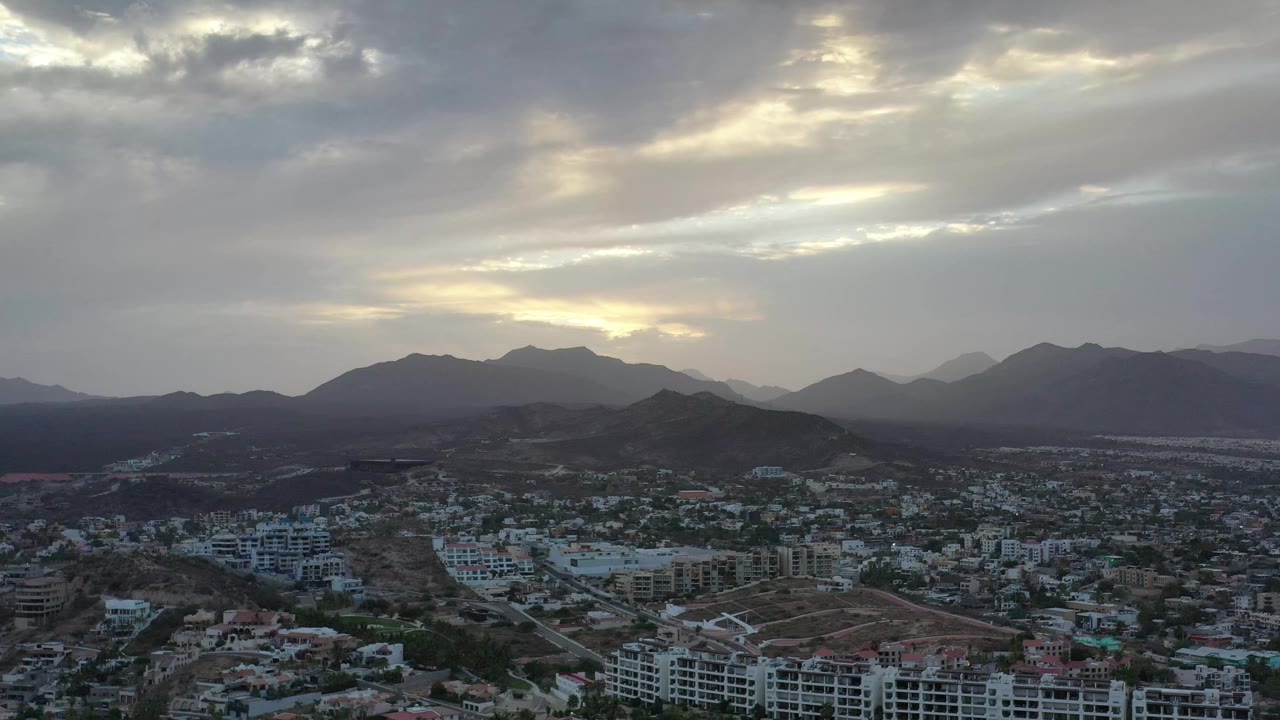 Los Cabos city blocks sit in the lower frame while mountain silhouettes and pale sunlight breaking through clouds dominate the background, with no visible people.