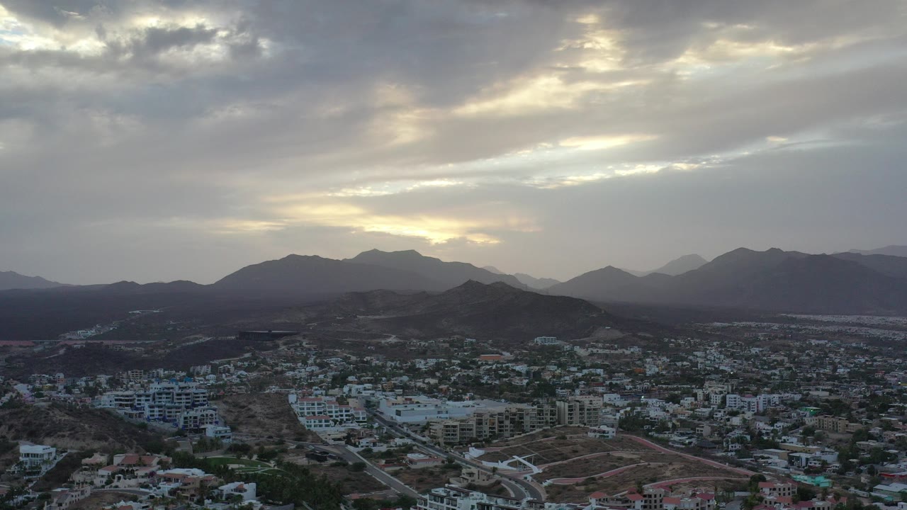 Wide aerial view of Los Cabos, Mexico, with clustered town buildings in the lower frame, dark hills and mountains beyond, broken gray clouds, and no visible people.
