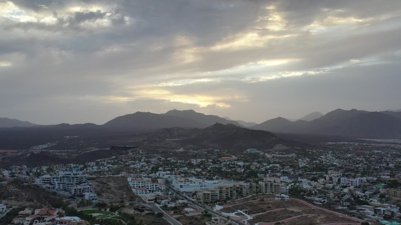 Opening aerial frame of Los Cabos with low-rise buildings and roads below dark mountain ridges, bright cloud breaks overhead, and no visible people.