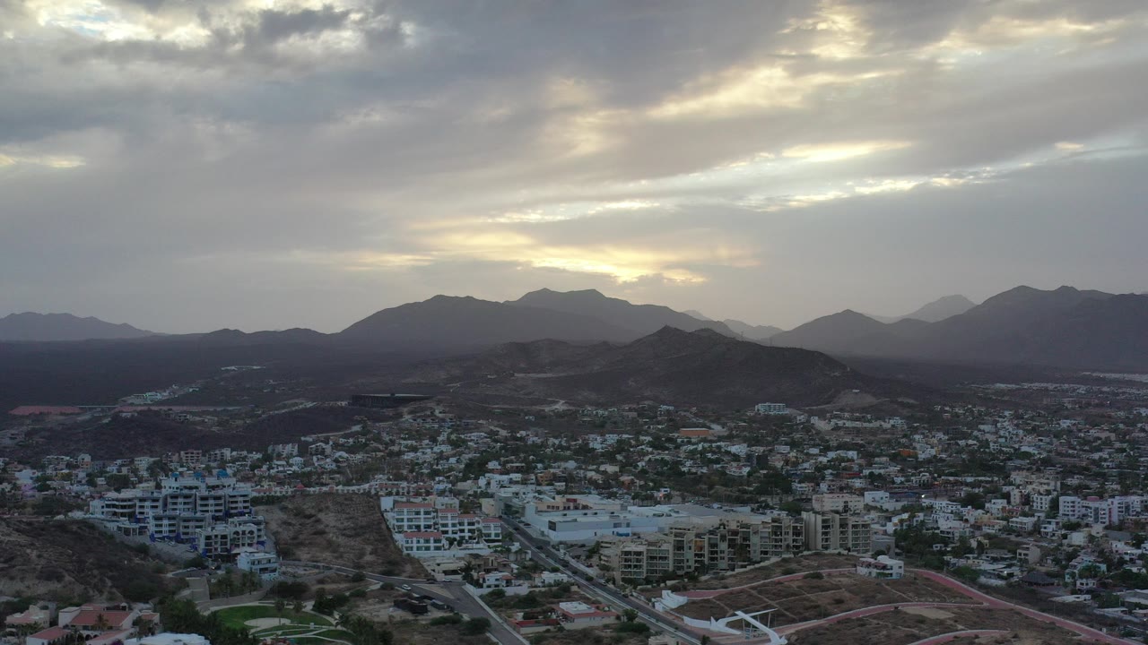Opening aerial frame over Los Cabos, Mexico, with residential buildings below, dark hills across the middle distance, layered mountains beyond, and no visible people.