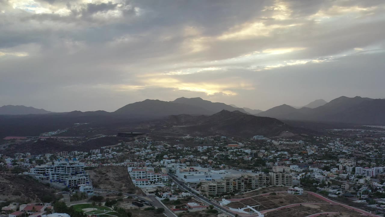 Los Cabos aerial view one second in, showing clustered houses and apartment blocks at the bottom of frame, a low ridge in the midground, distant mountains, and no visible people.