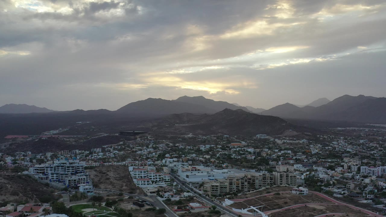 Aerial frame from Los Cabos with white and tan buildings in the foreground, a dark rounded hill near center, hazy mountains in the background, and no visible people.