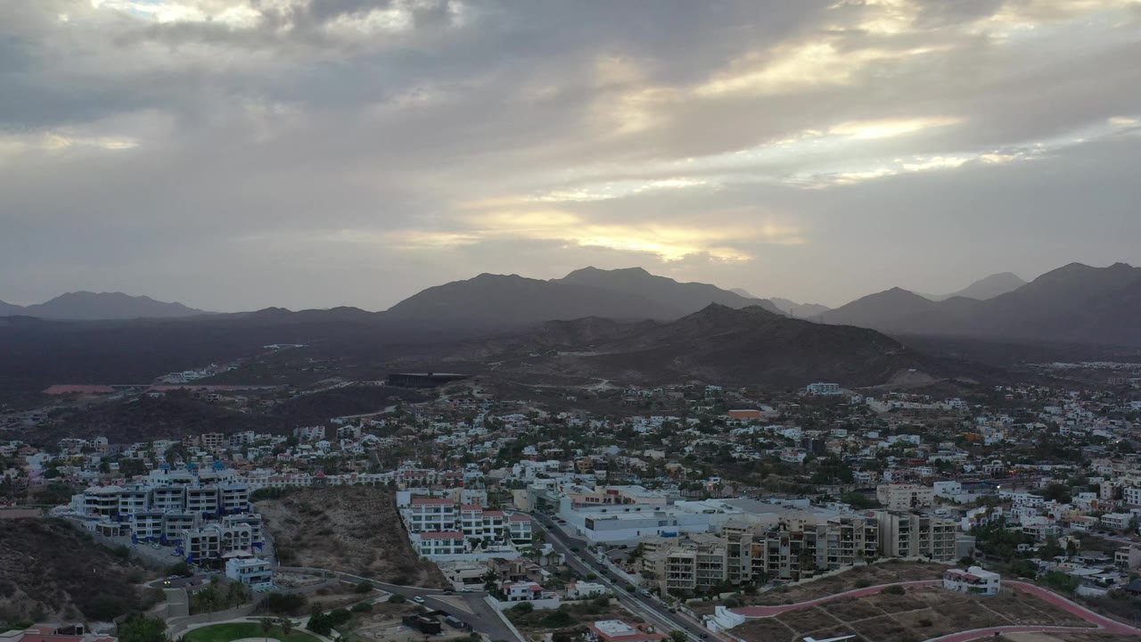 Los Cabos aerial frame with compact buildings and roads in the lower third, dark foothills across the center, distant mountain layers, cloud breaks, and no visible people.