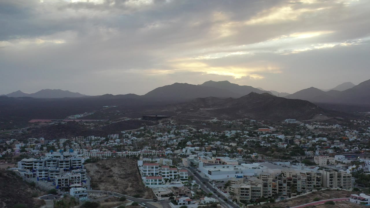 Aerial view of low-rise city buildings below dark hills and cloudy sky.
