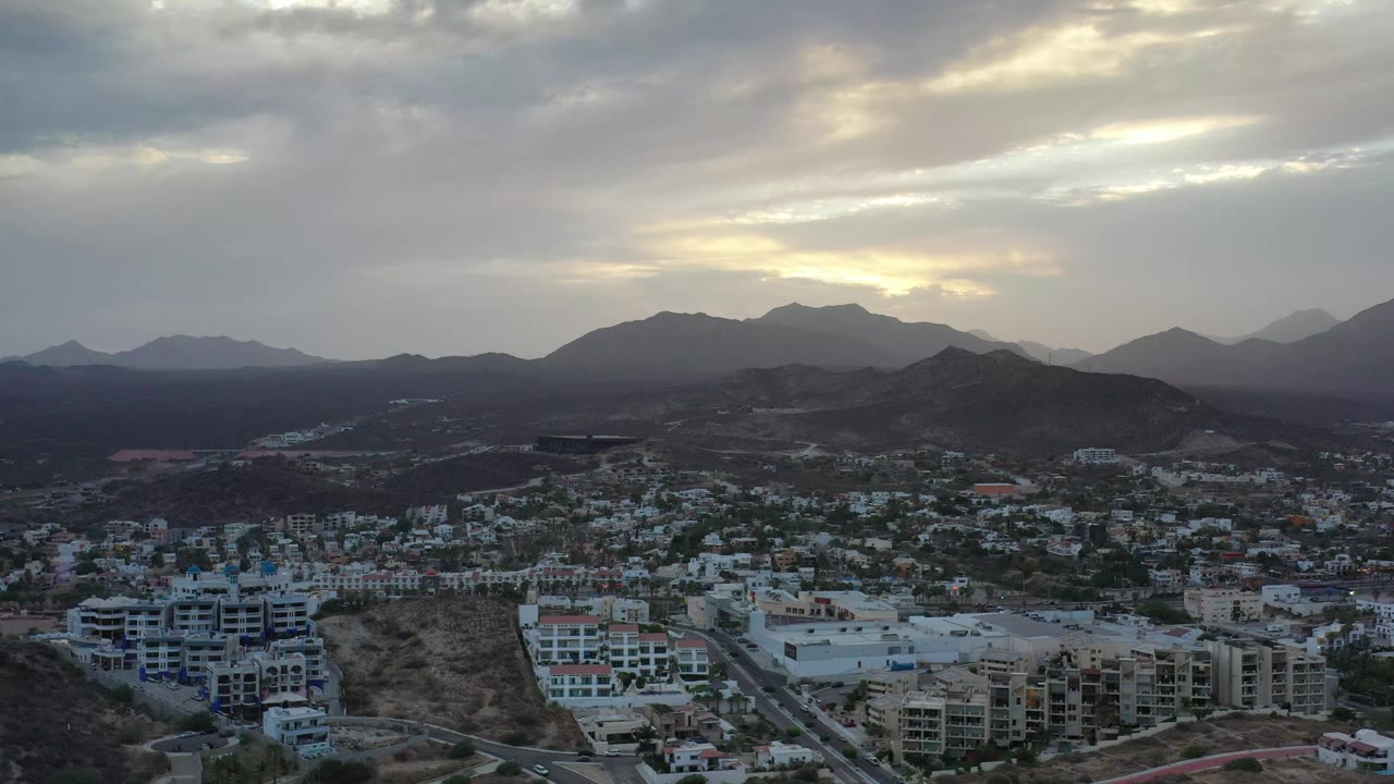 Aerial Los Cabos frame with white and tan apartment blocks, a road corridor, dark ridges, and a muted sky with brighter cloud openings, with no visible people.