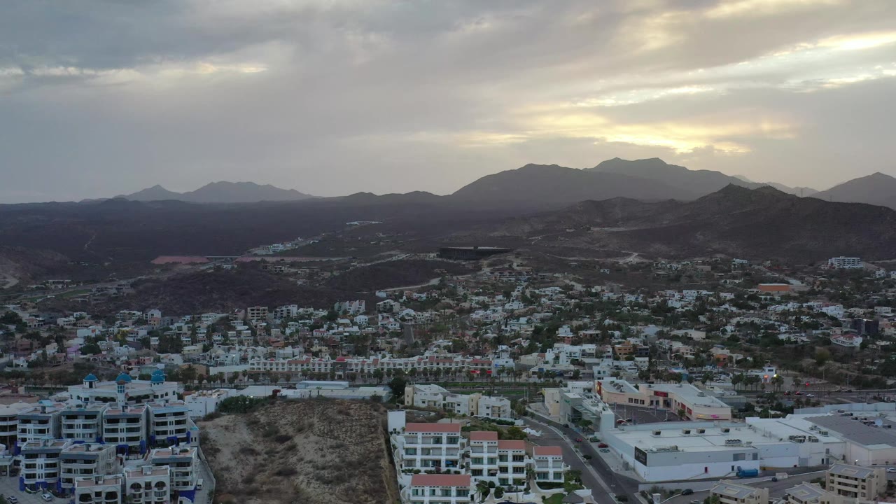 Wide Los Cabos, Mexico view with pale rooftops and residential streets in the lower frame, rugged hills across the center, layered mountains on the horizon, and no visible people.