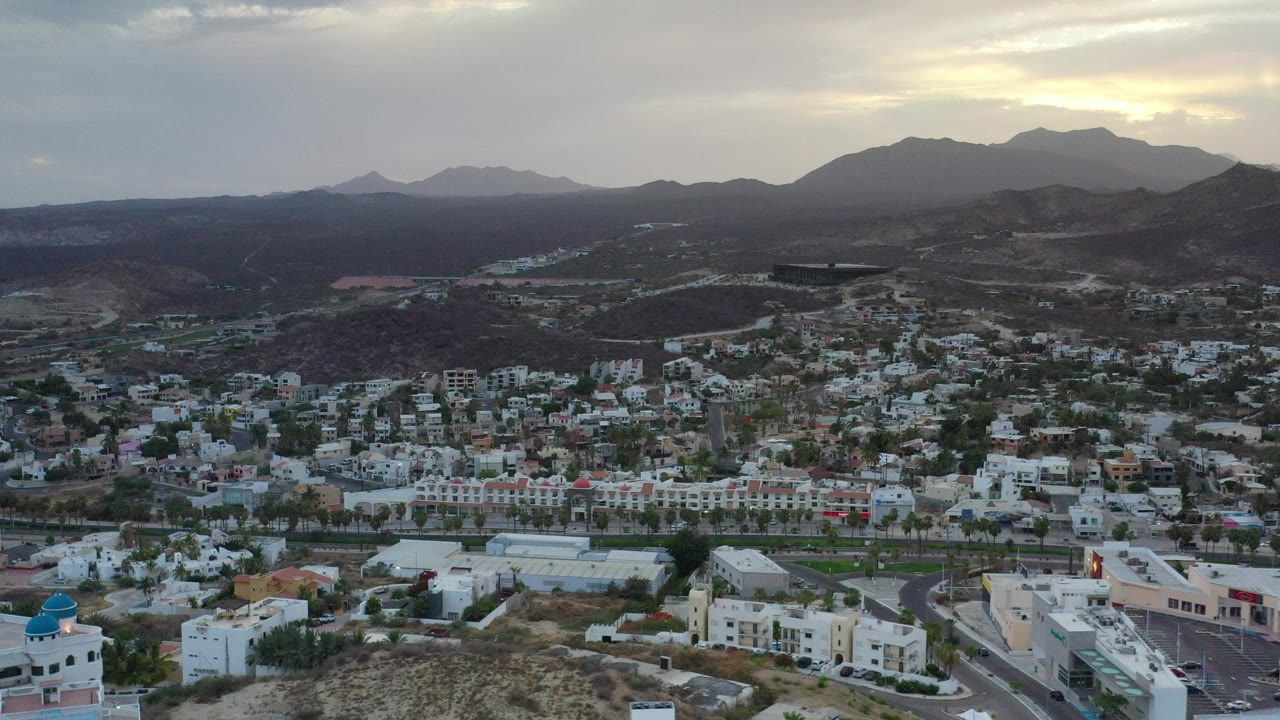 Wide aerial frame over a Los Cabos residential area with white homes, a road grid, palms, dark hills, muted sunset clouds, and no visible people.