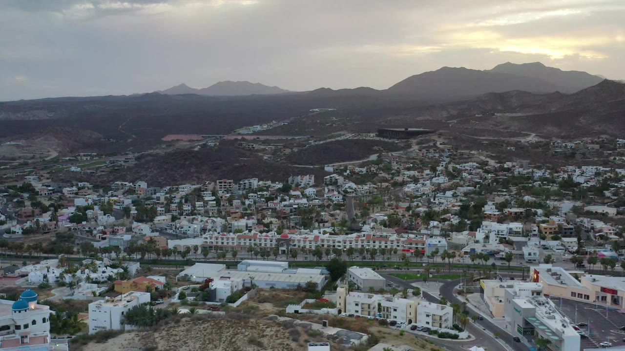 Elevated Los Cabos view with residential blocks, a curving road in the lower frame, scattered palms, dark mountain ridges, and no visible people.
