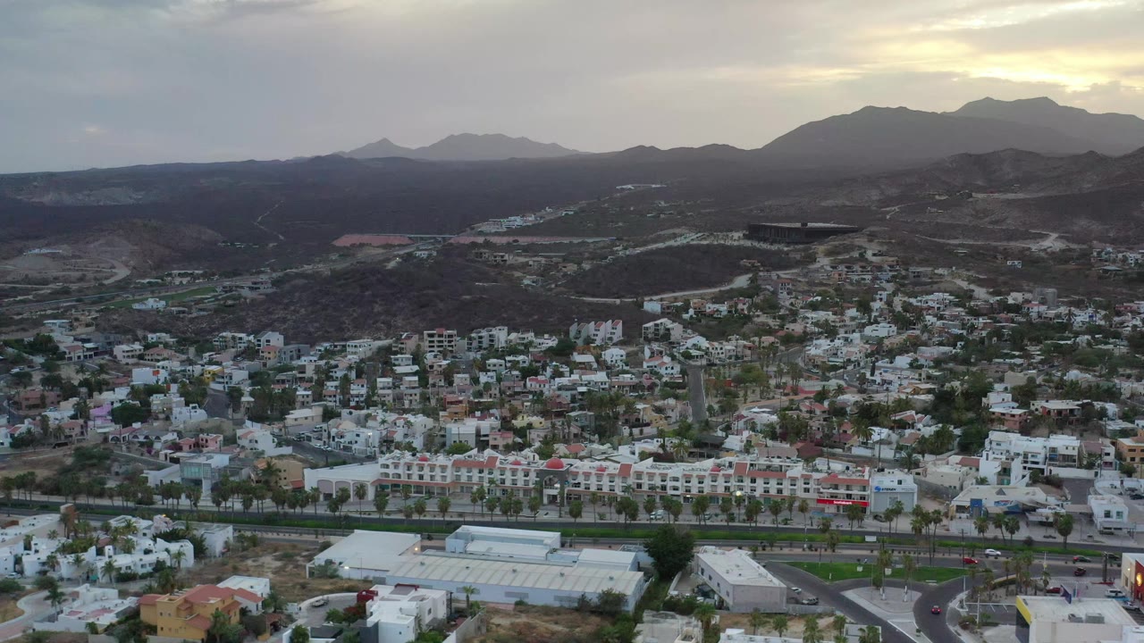Second frame holds on Los Cabos housing blocks, a road along the lower edge, dark hills beyond town, distant mountains, and no visible people.