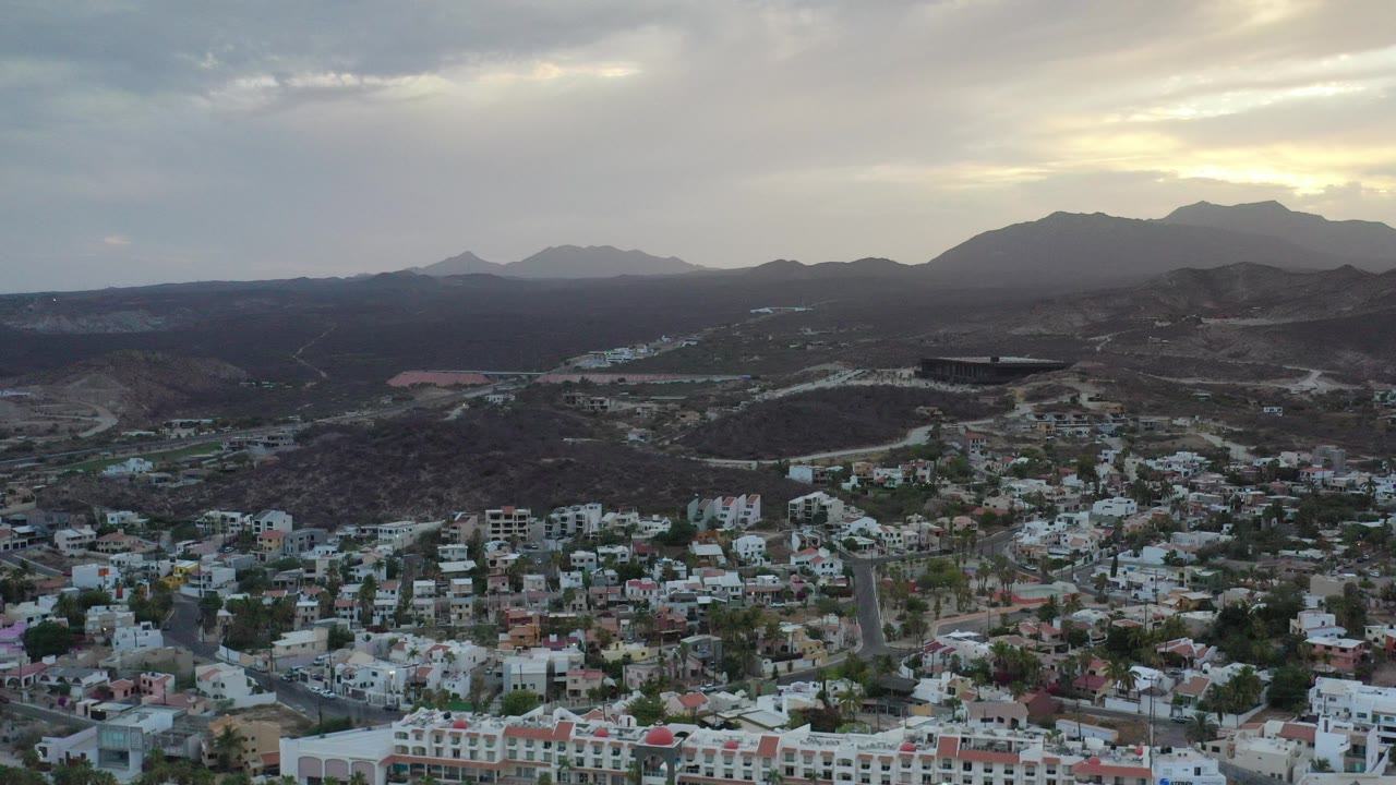 Aerial view of hillside town below cloudy mountains.