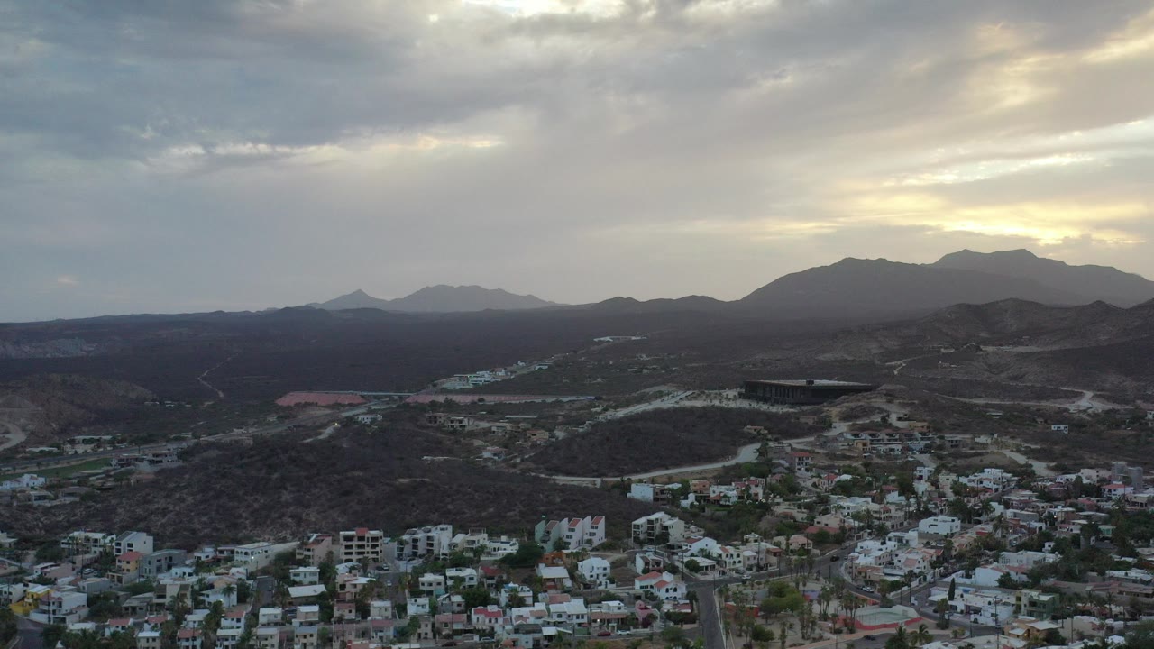 Opening aerial frame of Los Cabos, Mexico, with residential buildings at the bottom, dark hills across the middle, mountain ridges on the horizon, cloud-filtered light, and no visible people.
