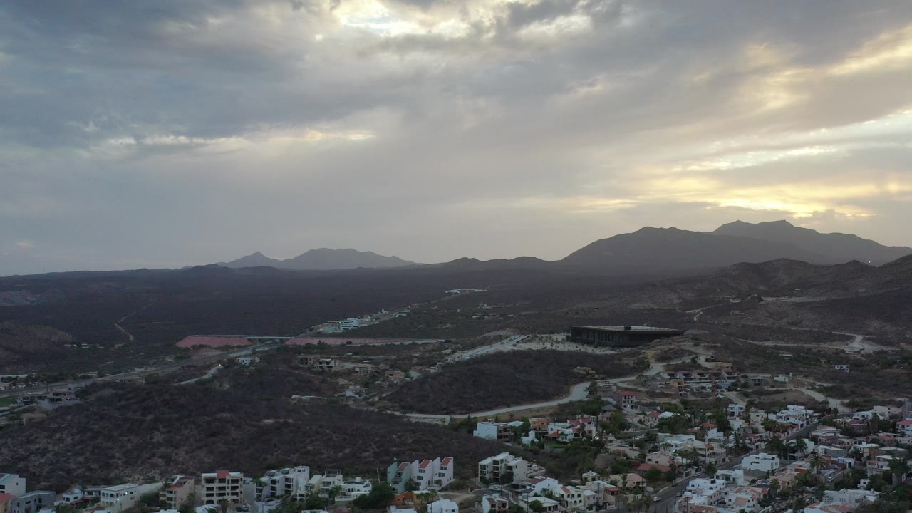 Los Cabos aerial at the opening moment, with hillside buildings along the lower edge, winding roads through dark terrain, distant mountains, layered clouds, and no visible people.