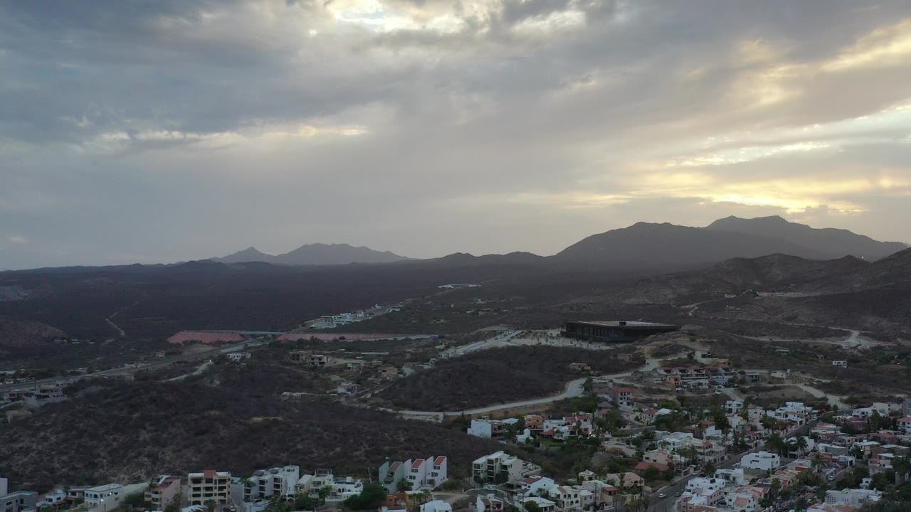 Aerial city edge with pale buildings, mountain ridges, and bright cloud breaks.
