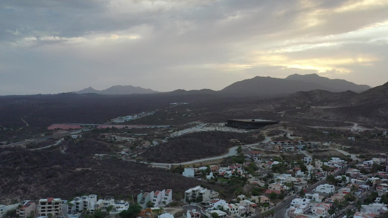 Aerial view of hillside homes, winding roads, and distant mountains under cloudy light.
