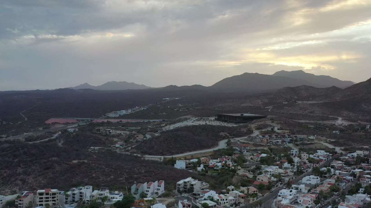 Aerial Los Cabos frame showing pale roads threading through brown hills, residential buildings along the lower edge, a dark rectangular structure in the midground, and no visible people.