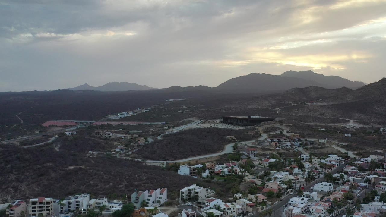Los Cabos hillside neighborhood with scattered buildings, dry slopes, and a cloudy mountain horizon.
