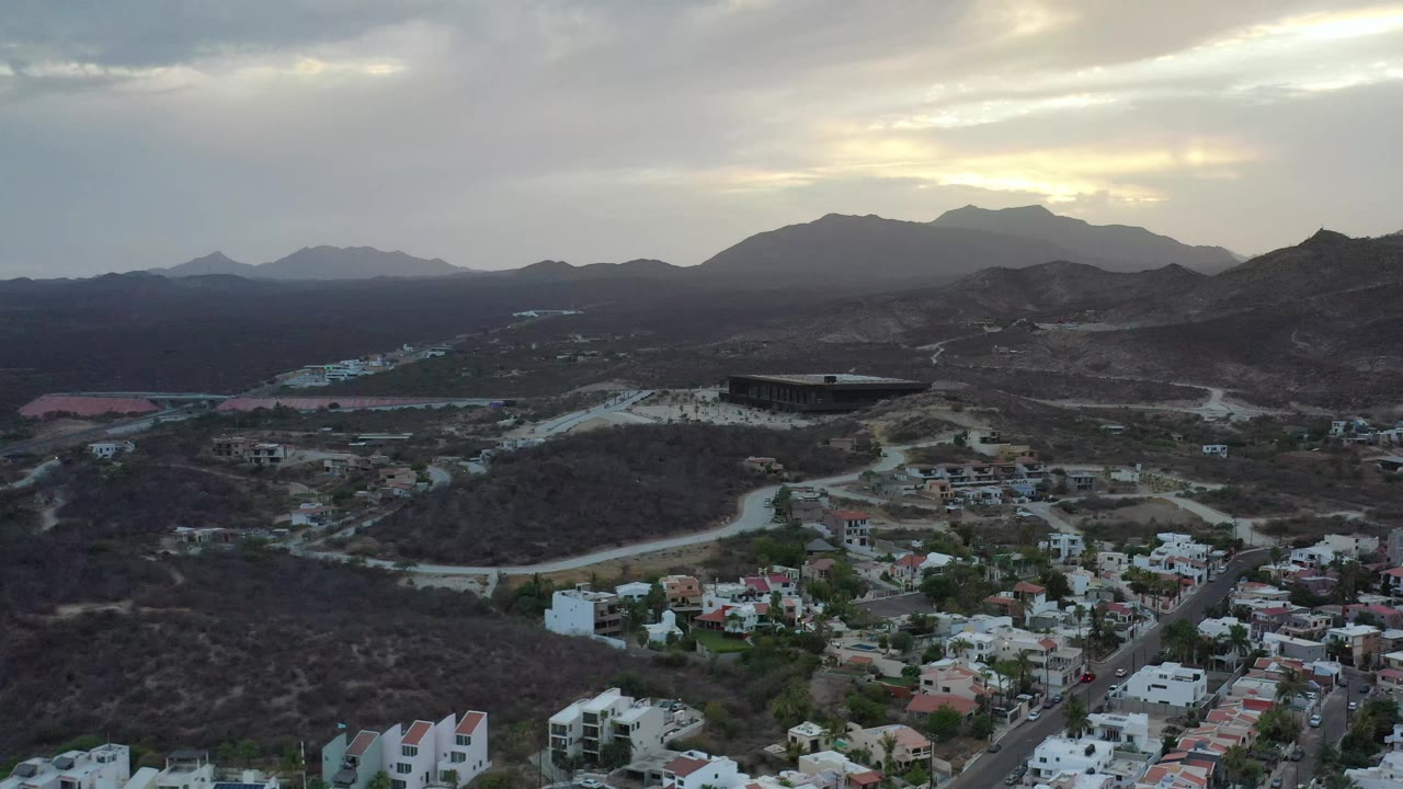 The aerial view over Los Cabos, Mexico holds on residential rooftops, a road curving through dark brush, the large central building, hazy mountains, cloud cover, and no visible people.