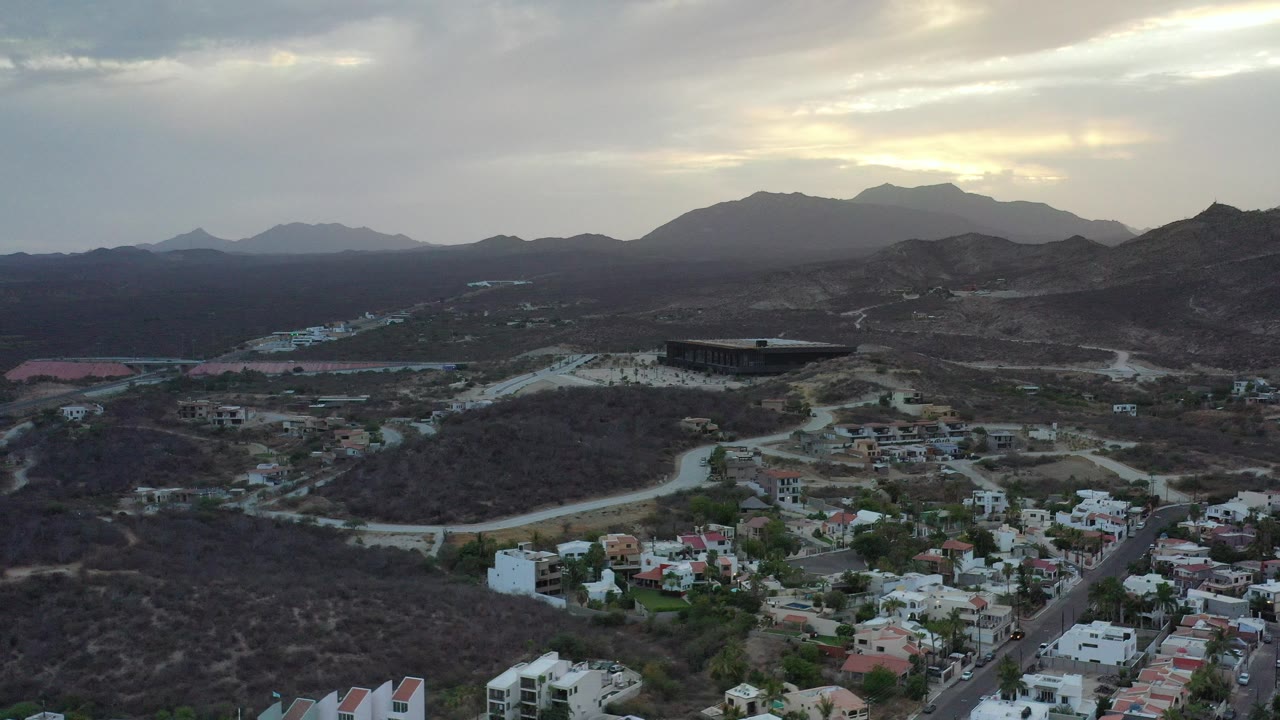 Aerial view of homes, roads, dry hills, and distant mountains under cloudy sunset light.