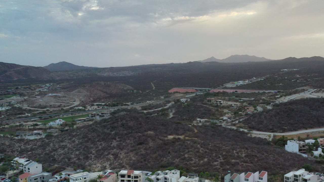 High aerial view in Los Cabos, Mexico, looking across dry hillside terrain, scattered white buildings, winding roads, distant mountains, and a cloudy sky, with no visible people.