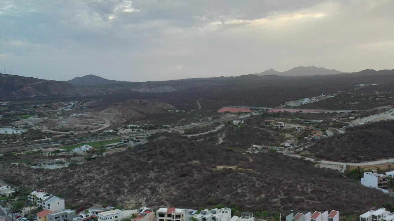 Wide aerial view of hills and town structures with mountains beneath a dim sky.