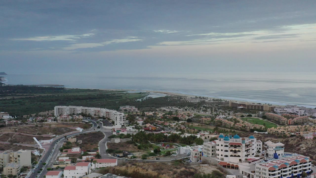 Aerial view of coastal town, roads, buildings, beach, and ocean under a clouded sky.