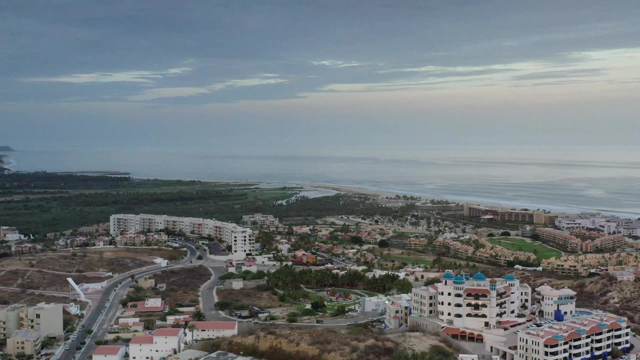 Second frame of the Los Cabos aerial view shows the same coastal neighborhood with white apartment buildings, road curves, darker vegetation near the shore, and no visible people.