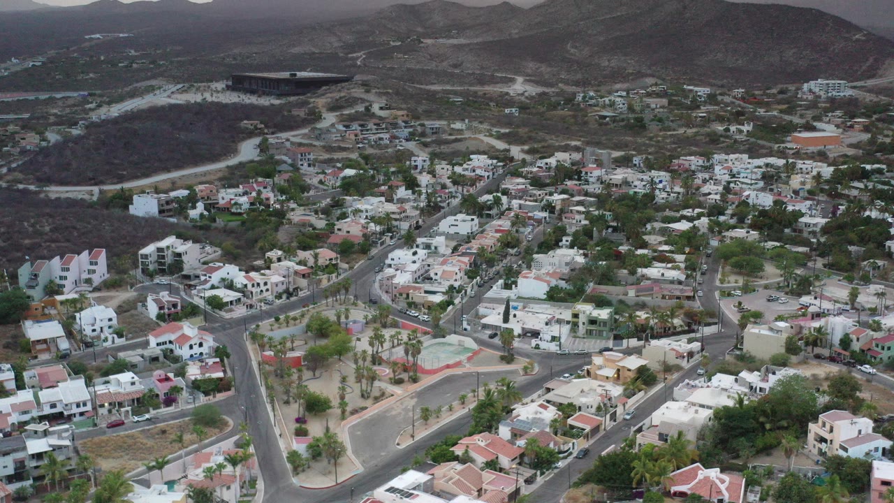 Opening aerial frame over Los Cabos, Mexico, with white homes, curving roads, dry hills, a distant water edge, and no visible people.