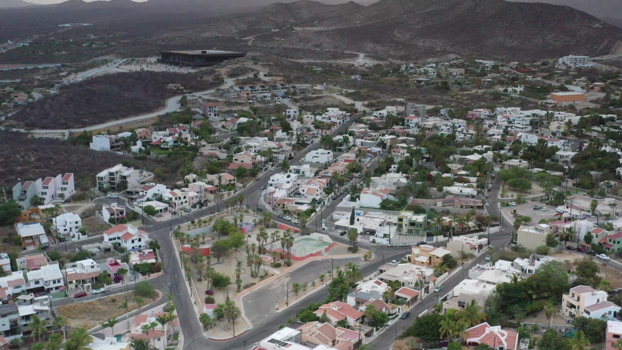 Los Cabos aerial view with a sloping residential grid, low white buildings, dark scrub-covered hills, and no visible people.