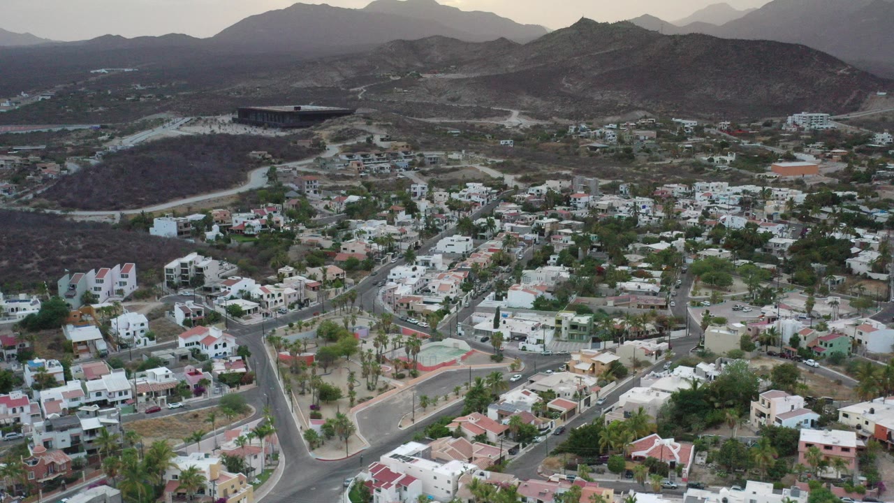 High aerial view over a Los Cabos residential area with white houses, curving roads, palms, dark hills, and hazy mountains beyond, with no visible people.