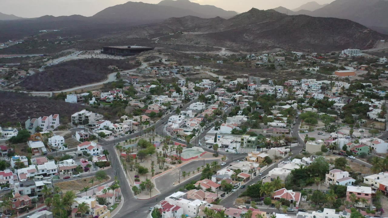 Wide drone view in Los Cabos showing dense low-rise homes, intersecting streets, small patches of trees, a dark hillside, and layered mountains in muted light, with no visible people.