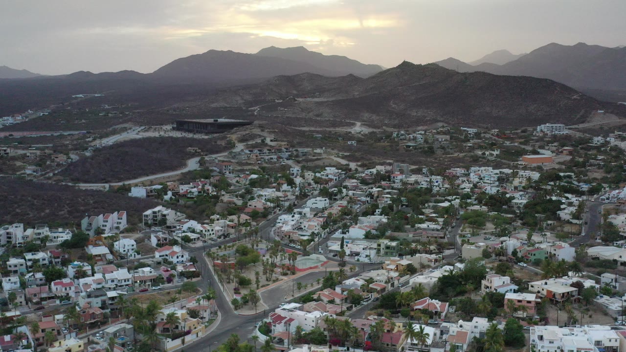Aerial view over Los Cabos, Mexico, showing hillside homes, winding roads, dark ridges, distant mountains, and no visible people at the start of the clip.