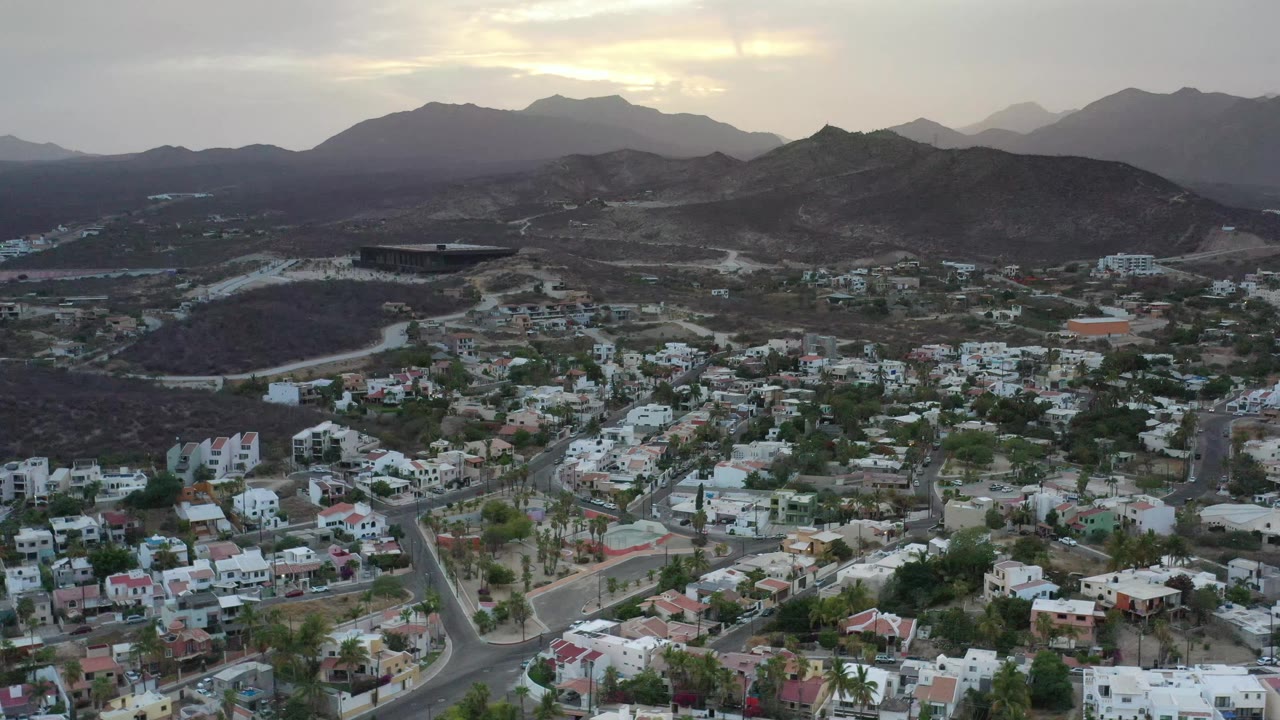 Elevated view of Los Cabos with clusters of white homes, curving roads, dry hills, hazy mountains, and no visible people one second into the clip.