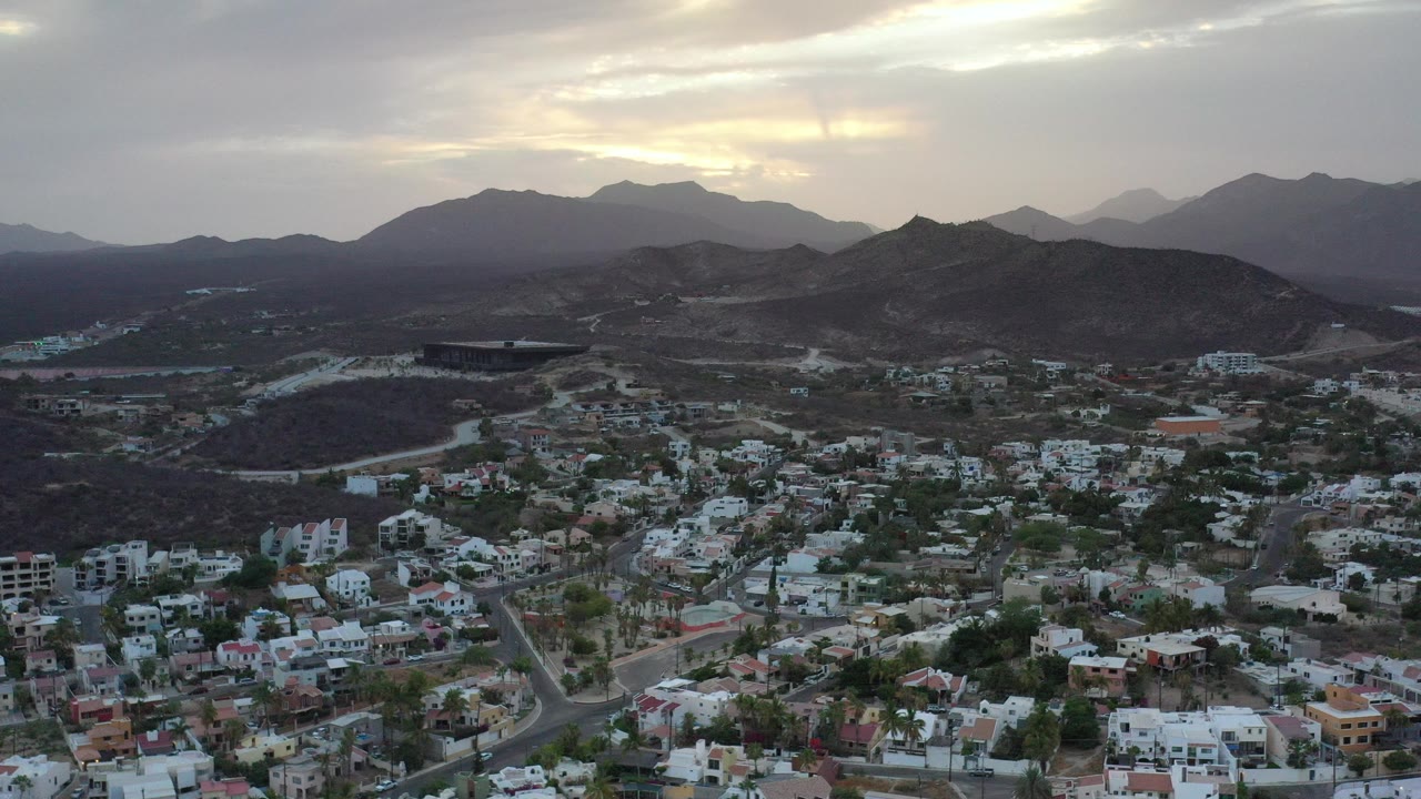 Wide aerial frame of Los Cabos, Mexico with low-rise buildings and roads in the lower foreground, dark hills and layered mountains behind, warm light breaking through clouds, and no visible people.