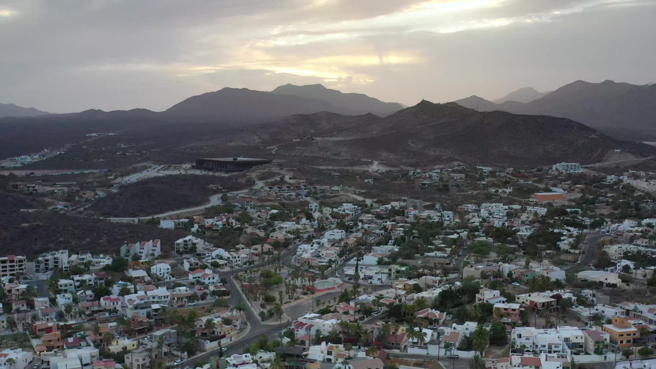 Second frame of Los Cabos, Mexico showing more of the residential grid below, a winding road near the hills, muted gray cloud cover, and no visible people.
